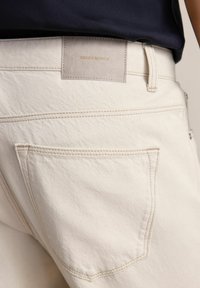 Białe dżinsy z widoczną tylną kieszenią, skórzaną metką z napisem "DENIM SUPPLY" oraz kontrastowym przeszyciem.