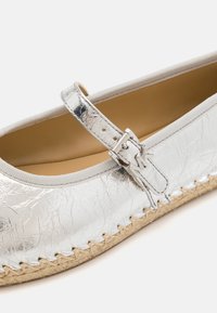 Gros plan sur une chaussure plate Mary Jane argentée avec une bride texturée et une semelle en jute tressée.