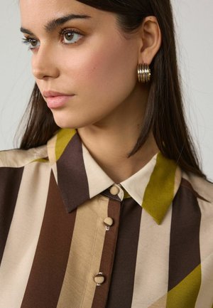 Junge Frau mit braunen Haaren trägt goldene Creolen und ein gestreiftes Hemd mit Knöpfen in Brauntönen, Beige und Olivgrün.