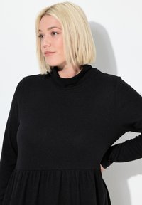 Sort langærmet kjole med høj hals, der har en tætsiddende overdel og en flagrende bund. Fremstillet af blødt stof med en glat tekstur.