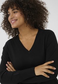 Black V-neck sweater with long sleeves, made of a smooth fabric. Model has curly hair and arms crossed in front of her torso.