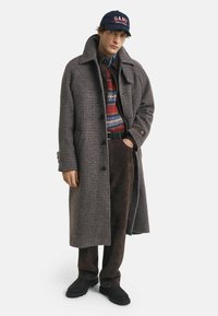 Man in een houndstooth lange jas, kleurrijke gebreide trui, bruine broek, zwarte schoenen en een marineblauwe pet met een rood logo.