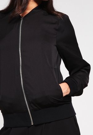 Femme portant une veste bomber noire zippée avec poignets et ourlet côtelés, une main dans la poche, sur un fond clair uni.