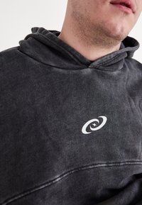 Tumši pelēks kapuce ar mīkstu tekstūru, ar baltu logo uz krūšu. Dizainā ir iekļauta ar stīgu regulējama kapuce un brīva piegriezuma forma.