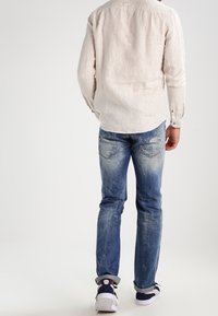Camisa de lino de color claro y mangas largas, vaqueros de mezclilla azul desgastados y zapatillas marinas con suelas blancas. Texturas de las telas visibles; diseño de corte casual.