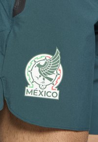 Πράσινα αθλητικά σορτς με λευκό έμβλημα που απεικονίζει ένα στυλιζαρισμένο ποδοσφαιρικό μπάλα με φτερά και το κείμενο "MÉXICO" σε πράσινες, κόκκινες και λευκές αποχρώσεις.