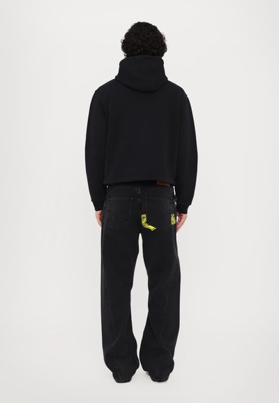 Un hoodie noir court avec une capuche épaisse, associé à un jean noir ample présentant un détail de conception en jaune néon sur la poche arrière.