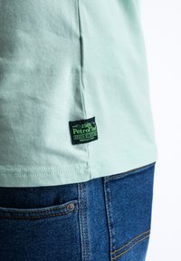 Camiseta de algodón verde menta con costura lateral y etiqueta negra con texto verde. Llevada con jeans de mezclilla oscura que muestran el detalle del bolsillo.
