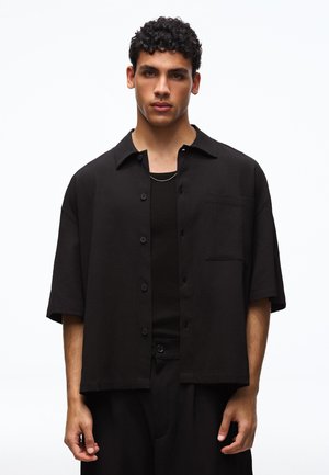 Fiatal férfi göndör sötét hajjal fekete rövid ujjú, gombos inget visel fekete trikó felett fehér háttér előtt.