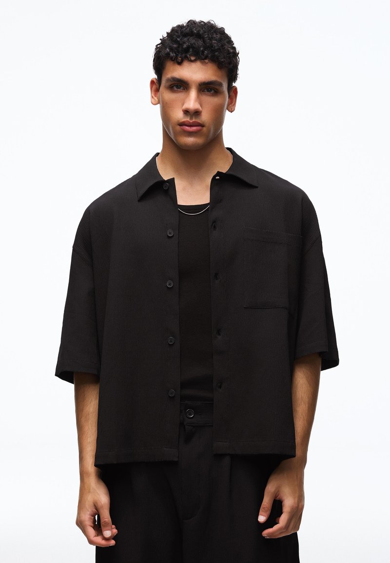 Jeune homme aux cheveux bouclés et foncés portant une chemise noire à manches courtes boutonnée sur un débardeur noir sur fond blanc.