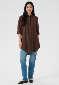 Rjava srajca s gumbi, z žepom, zavihanimi rokavi in zaobljenim robom, združena s svetlo modrimi kavbojkami s pravimi hlačnicami in črnimi balerinkami.