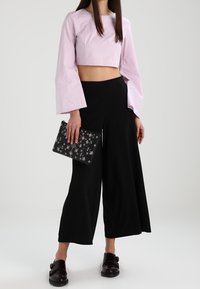 Top corto de color rosa claro con mangas acampanadas, pantalones de pierna ancha negros, sosteniendo un clutch negro con estampado y usando zapatos negros sin cordones.