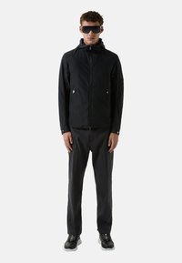 Zwarte lichtgewicht jas met capuchon, voorzien van ritszakken en een gladde textuur, gecombineerd met donkere broek en zwarte sportschoenen.
