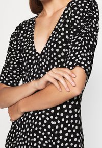 Žena v černých šatech s bílými nepravidelnými puntíky, s výstřihem do V a nařasenými krátkými rukávy, s crossed arms a přirozenými nehty.