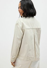Beige denim jack met verticale rode strepen, voorzien van een plooien aan de achterkant en knoopdetails bij de manchetten. Gestructureerde stof met een casual pasvorm.