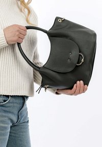 Tummanvihreä nahkakäsilaukku, jossa on teksturoitu pinta, kaareva kahva ja kultaisia metalliyksityiskohtia. Hän pitää laukusta, jonka päällä on vaaleankermanvärinen neule.