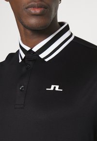 Svart polo-tröja med en texturerad yta, med en vit randig krage och en liten vit logotyp på bröstet. Fyra knappslå.