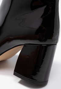 Black patent leather ankle boot featuring a square, chunky heel and a smooth texture. Interior shows a beige lining.