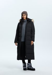 Musta vuorellinen takki, jossa on huppu ja keinokarvasauma, päällä harmaa poolopaita ja mustat housut. Harmaat ja mustat karvavuorelliset saappaat.