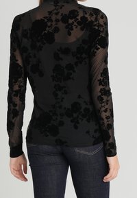 Svart långärmad topp med genomskinligt tyg och ett strukturerat blommönster. Strukturerade detaljer står i kontrast mot en solidd svart bas.