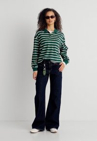 Γυναίκα με κατσαρά μαλλιά, που φοράει πράσινο και λευκό ριγέ πόλο με μακριά μανίκια, σκούρα τζιν φαρδιάς γραμμής, λευκά παπούτσια και γυαλιά ηλίου, στέκεται μπροστά σε λευκό τοίχο.