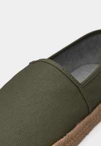 Πράσινο ελιά καμβά παπούτσι χωρίς κορδόνια με καφέ πλεγμένη σόλα από τζούτ και ορατή ραφή κατά μήκος του άνω άκρου.