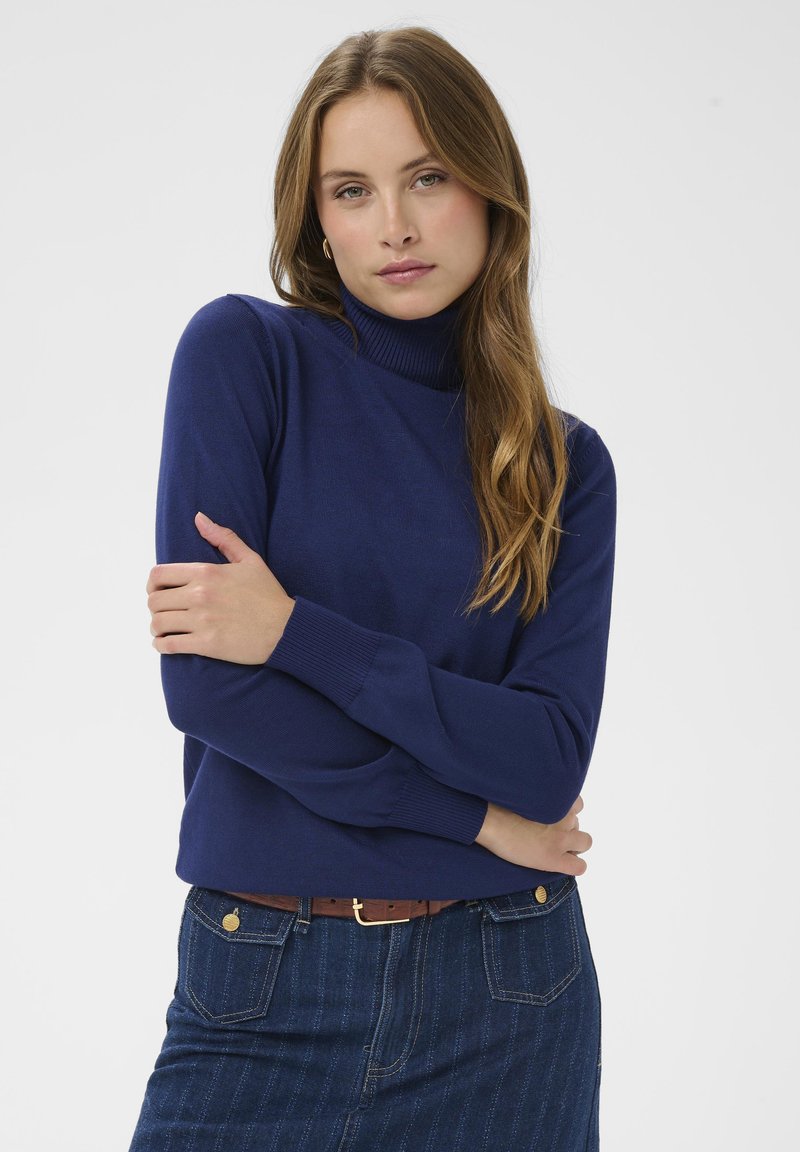 Pull à col roulé bleu marine en maille douce, à manches longues et poignets côtelés, associé à une jupe en denim à rayures verticales.