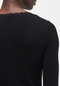 Schwarzes, geripptes Longsleeve-Oberteil mit einem gewellten Halsausschnitt. Die Stoffstruktur ist glatt mit vertikalen Streifen.