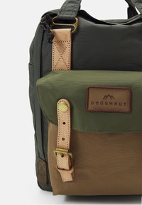 Sac à dos en tissu vert et marron avec des accents en cuir, doté d'une boucle métallique et d'un patch logo. Surface textur ée et design structuré.