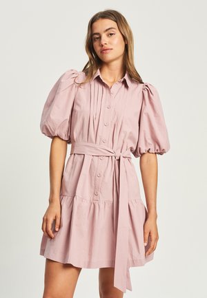 Junge Frau trägt ein hellrosa Hemdkleid mit Puffärmeln und einem gebundenen Taillengürtel, steht vor einem einfarbigen hellen Hintergrund.