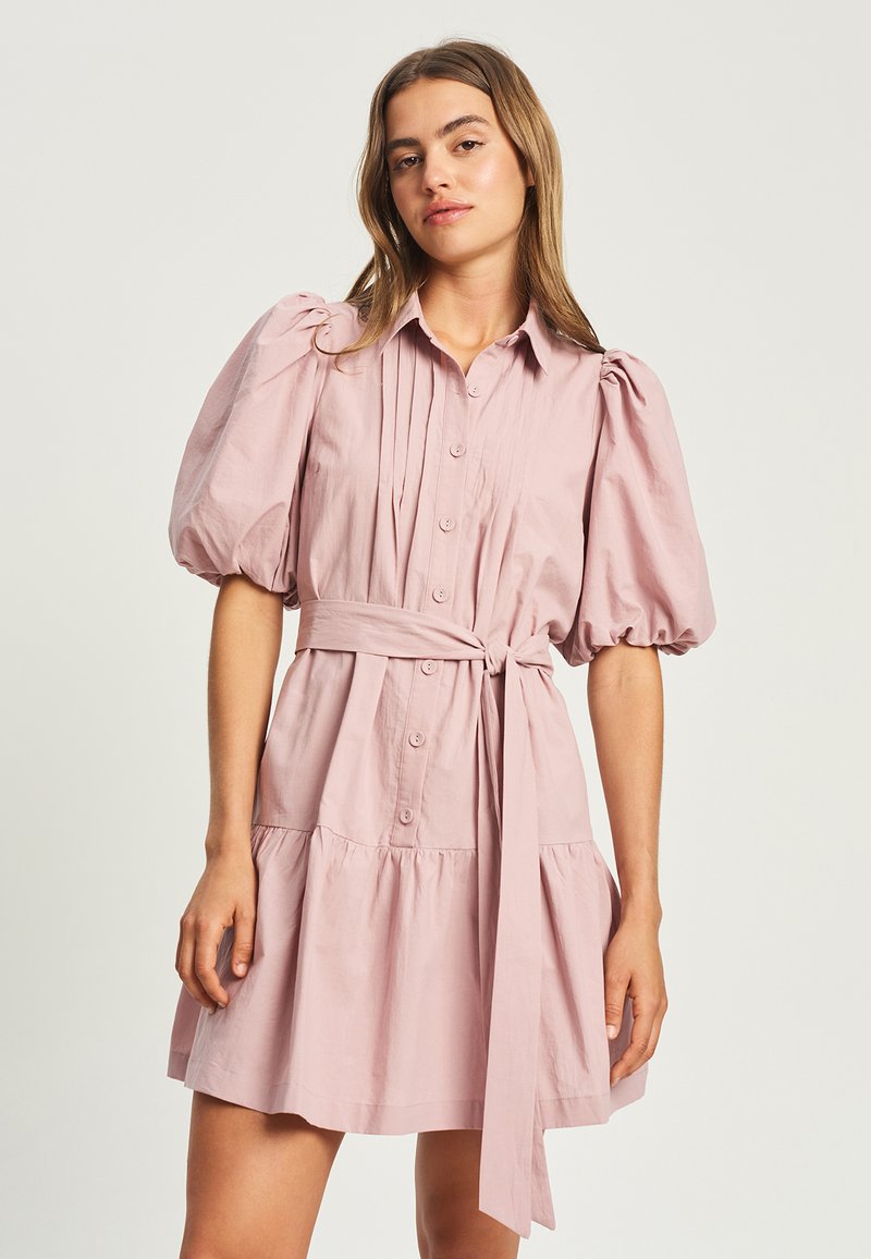 Ung kvinde iført en lyserød knap-op kjole med pufærmer og bælte bundet i taljen, stående mod en ensfarvet lys baggrund.