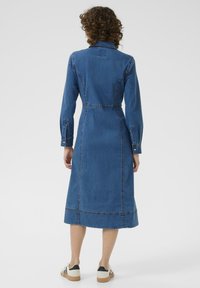 Rochie din denim cu mâneci lungi, guler, detalii cusute și lungime midi. Purtați cu încălțăminte casual, evidențiind o croială lejeră.