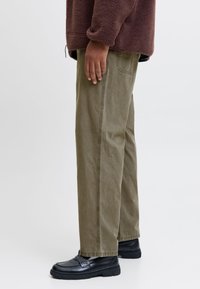 Olijfgroene katoenen broek met een rechte snit en zijnaaddetails, gecombineerd met zwarte matte schoenen. Warme bruine fleece bovenkleding zichtbaar boven de taille.