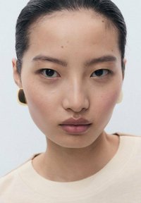 Jeune personne à la peau lisse, portant un maquillage minimal, vêtue d'un haut beige et de grandes boucles d'oreilles carrées dorées et noires, regardant directement devant elle.