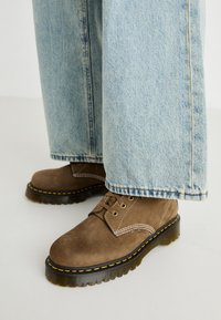 Botas de ante marrón con cordones y costuras amarillas, con suelas de goma gruesas; llevadas con unos pantalones vaqueros de pierna ancha de color azul claro, con dobladillo deshilachado.