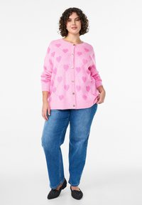 Roosa kardigan suurte südamekujunditega, nööbiga kinnitatav ja soonikkoes varrukasäärtega. Kandke seda koos siniste teksade ja mustade madalate kingadega vabaaja stiili jaoks.