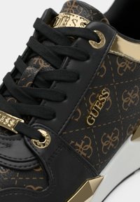 Sneaker nera con una trama testurizzata e dettagli dorati e di branding. Include lacci neri e un dettaglio metallico vicino alla suola.