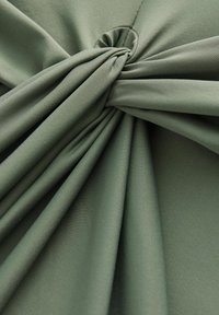 Grüner Stoff mit einem leichten Glanz, der sanfte Drapierungen und ein geknotetes Detail in der Mitte aufweist, und eine glatte, strukturierte Oberfläche zeigt.