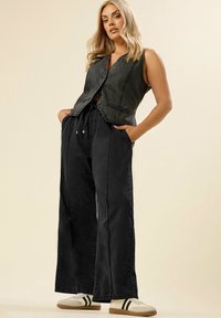 Femme portant un gilet en denim noir sans manches, un pantalon noir à jambes larges avec ceinture à cordon, des baskets blanches avec des rayures noires, posant avec les mains dans les poches.