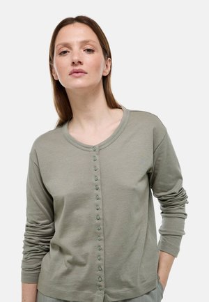 Femme aux cheveux bruns lisses portant une chemise boutonnée à manches longues couleur vert olive, une main dans la poche, sur fond gris.