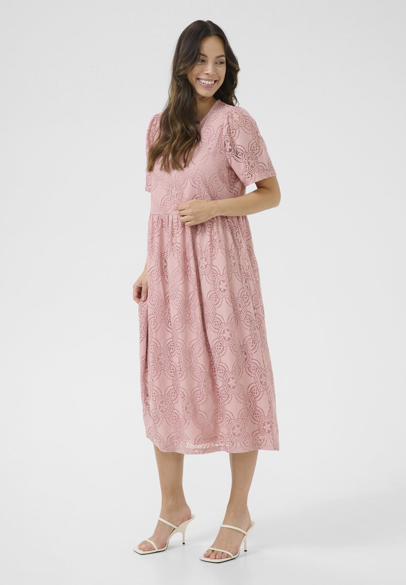 Rosa spets midi-klänning med korta ärmar, blommönster, rynkad midja och en flödande siluett. Matchad med vita klacksandaler.