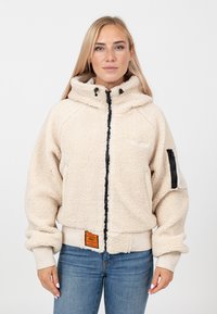 Kremowa polarowa kurtka z wysokim kołnierzem, czarnym zamkiem i boczną kieszenią. Posiada ściągacze przy mankietach oraz naszywkę z logo na wysokości talii.