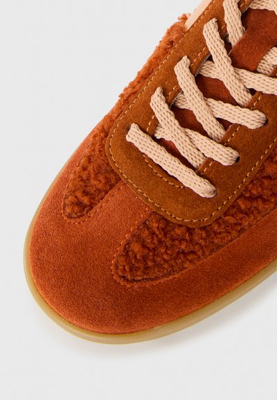 Sneaker in arancione ruggine con punta, lacci beige, caratterizzato da pannelli in suede misto e tessuto testurizzato, su uno sfondo chiaro.