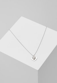 Collier chaîne en argent avec deux pendentifs en forme de cœur, l'un uni et l'autre incrusté de petites pierres claires, présenté sur un cube blanc.