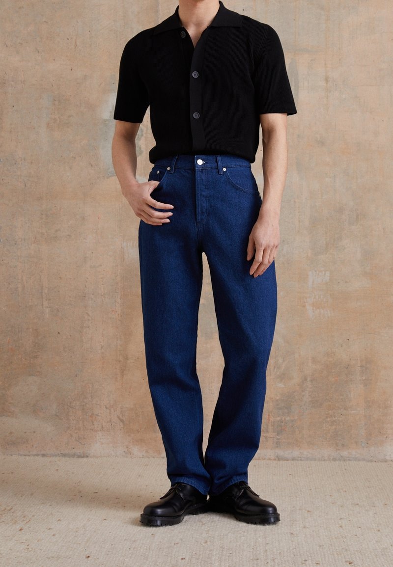 Personne portant une chemise noire à manches courtes boutonnée, un jean bleu foncé taille haute et des chaussures noires à lacets, debout contre un mur beige.