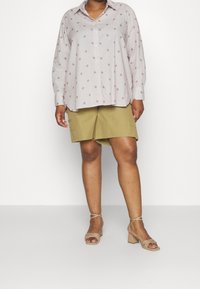 Camicia a maniche lunghe di un rosa chiaro con piccoli motivi geometrici rossi, abbinata a pantaloni corti beige a ginocchio e sandali con tacco blocco beige.