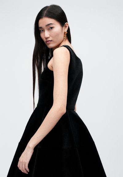 Femme aux longs cheveux noirs portant une robe noire sans manches et des boucles d'oreilles créoles torsadées dorées, debout devant un fond clair uni.