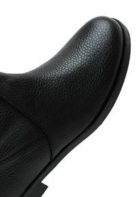 Bottine en cuir noir présentant un bout arrondi, une surface texturée et une semelle en caoutchouc basse. Détail de couture visible sur le côté.