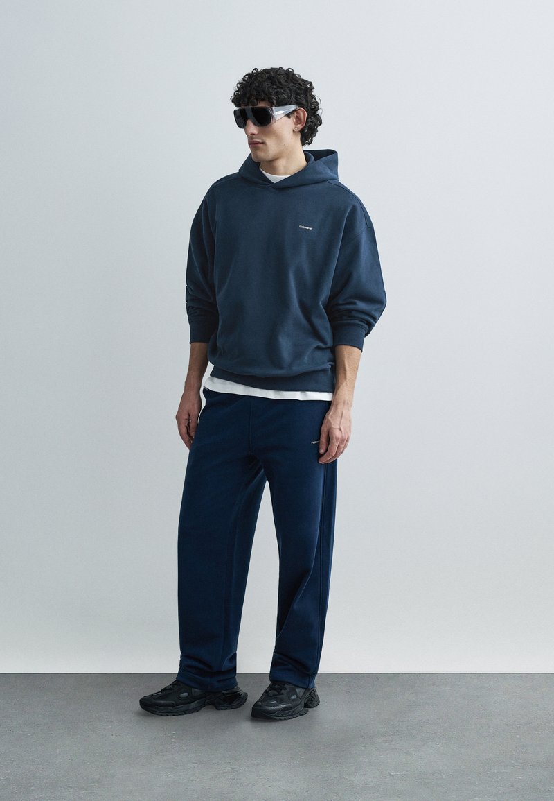 Navy blue hooded sweatshirt with a relaxed fit, paired with matching jogger pants. Black sneakers and oversized black sunglasses complete the look.