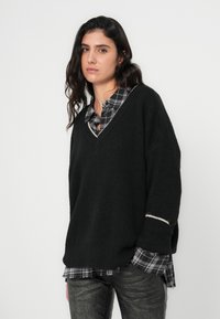 Melnas adītas džemperis ar dziļu V-veida izgriezumu, kam ir teksturēts pārklājums un baltas uzsveres uz piedurknēm, valkāts virs melnas un pelēkas pleda krekla.